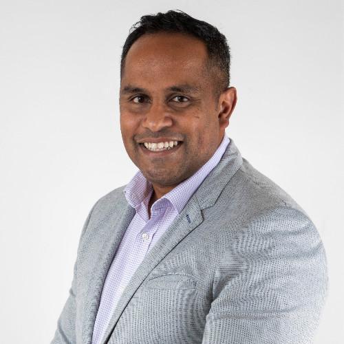Image of Rajiv wearing a grey suit jacket and lavender shirt in front of a grey background. He is smiling at the camera.